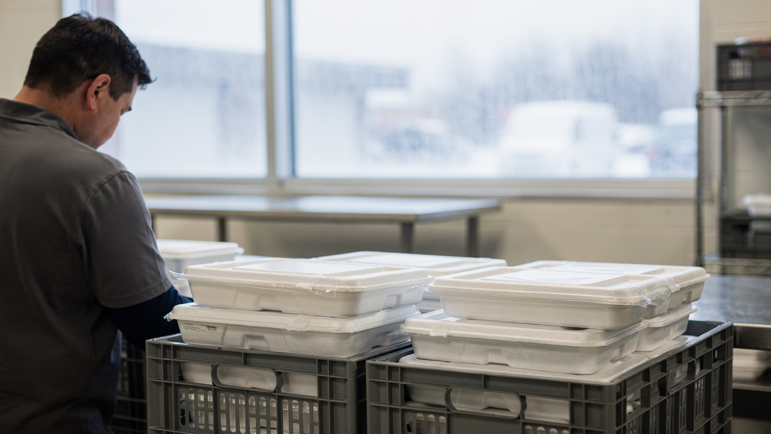 A kitchen worker packs sealed foam meal containers into grey plastic crates beside a frosted window with a snowy parking lot outside.