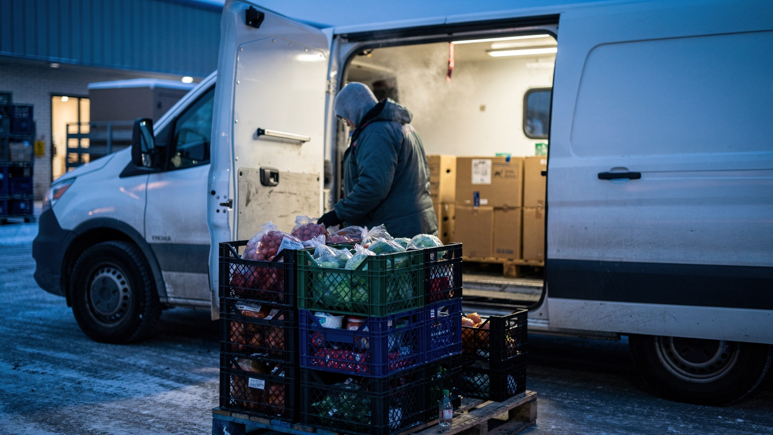 A hooded worker loads crates of vegetables and dairy into a refrigerated delivery van at a snowy grocery dock before dawn.