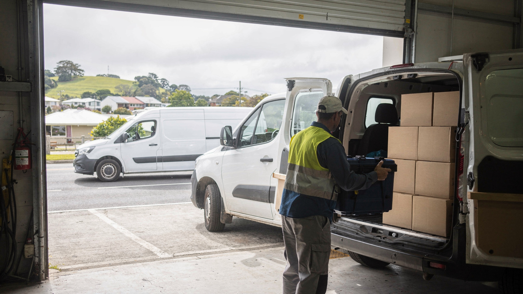 A delivery driver in a hi-vis vest loads an insulated tote into a white van stacked with plain cartons at an open depot roller door.