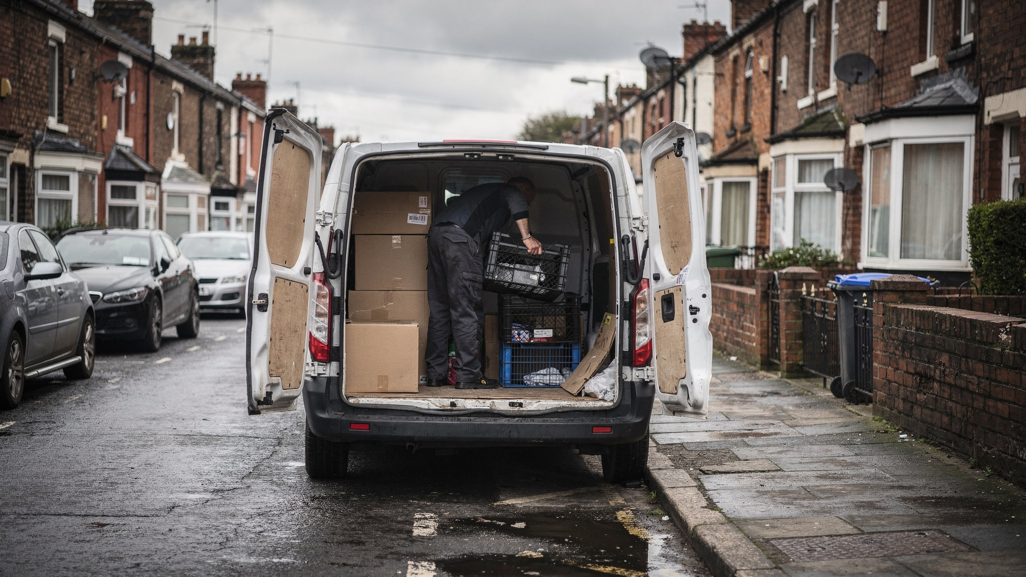 A white panel van with rear doors open on a wet terraced street as a driver lifts a crate of supplies from the cargo area.