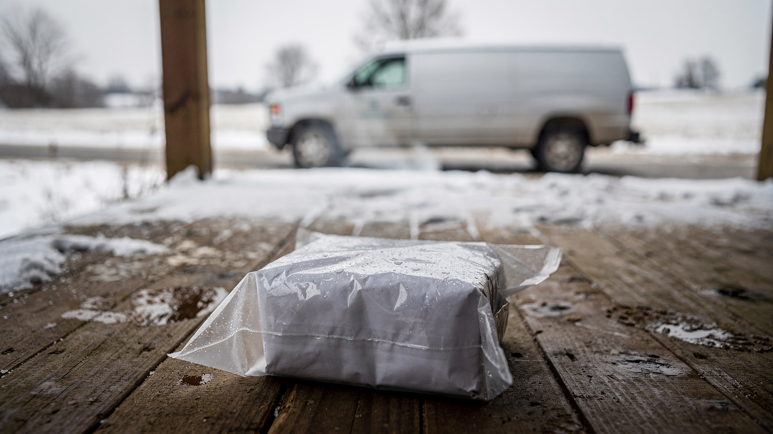 A parcel sealed in a wet plastic bag rests on a snowy wooden porch while a white delivery van waits on the drive beyond.